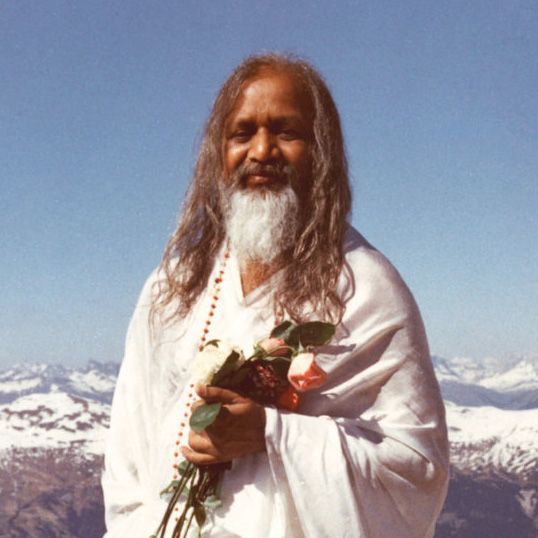 Maharishi Mahesh Yogi hält einen Blumenstrauß, im Hintergrund ist eine Berglandschaft