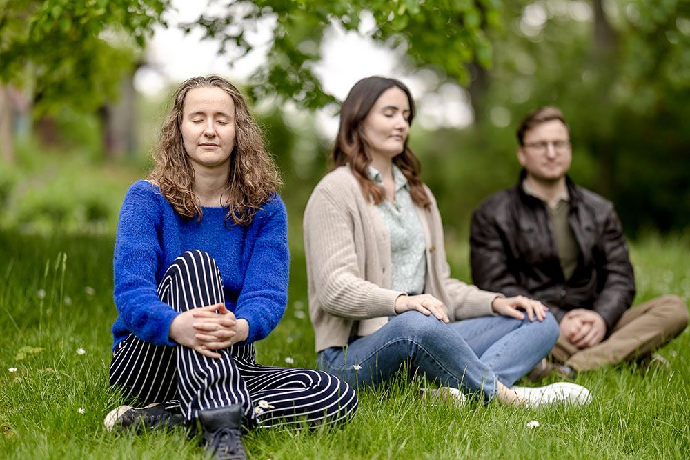 Drei junge Personen sitzen draußen auf einer Wiese mit geschlossenen Augen und meditieren.